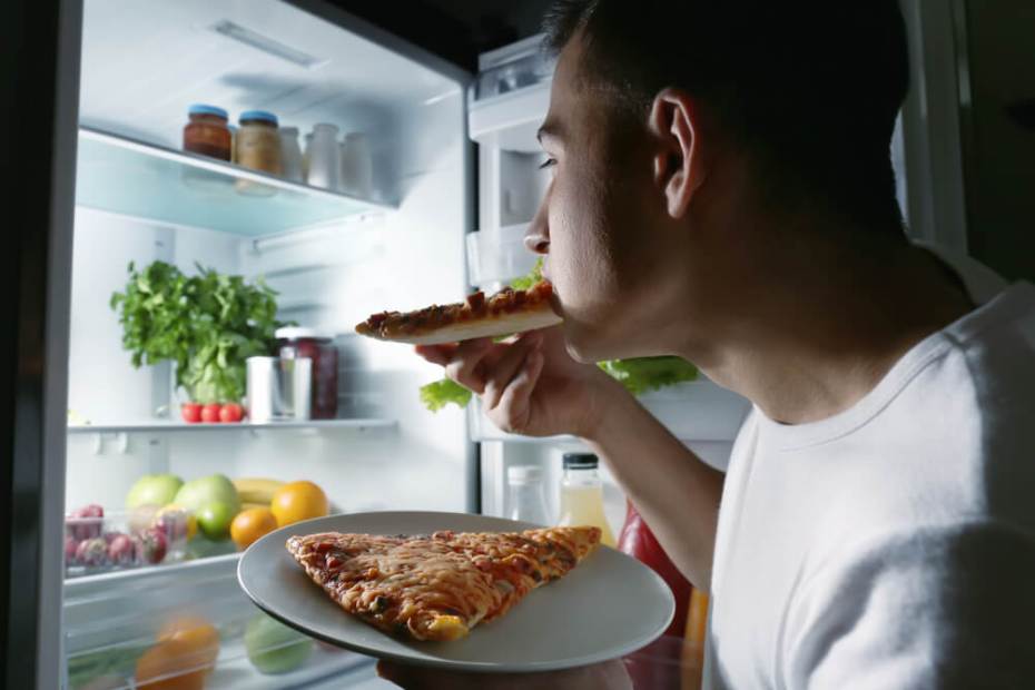 한 남자가 밤에 냉장고 문을 열고 피자를 먹고 있다